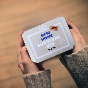 Może przedstawiać: Prostokątna, jasnoszara puszka z zaokrąglonymi rogami trzymana jest w dwóch dłoniach. Puszka zawiera tekst "NEW HOME SURVIVAL KIT IN A TIN" w kolorze niebieskim i białym, z małą grafiką domu. Etykieta głosi "PACKED WITH FUN".