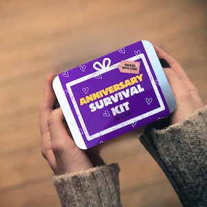 Puede incluir: Una lata rectangular morada con texto blanco que dice "ANNIVERSARY SURVIVAL KIT IN A TIN". La lata tiene un borde blanco y está decorada con pequeños corazones. Las palabras "PACKED WITH LOVE" están en una etiqueta rosa.