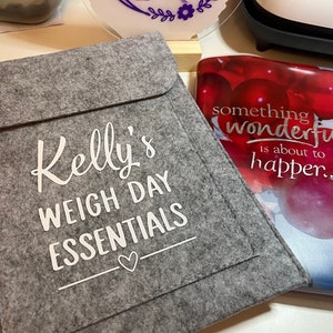 May include: A grey felt pouch with the text "Kelly's Weigh Day Essentials" in white lettering. A small heart is below the text. The pouch is lying on a table next to a book with the text "something wonderful is about to happen..." in white lettering on a red background.