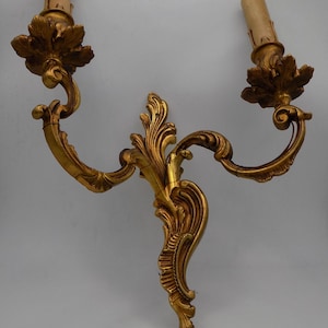 Può includere: Un'applique da parete in metallo dorato con due portacandele. L'applique ha un design decorativo e ornato con volute e fioriture intricate.