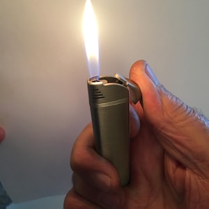 Puede incluir: Un encendedor plateado con una llama azul ardiendo. El encendedor está siendo sostenido en una mano.