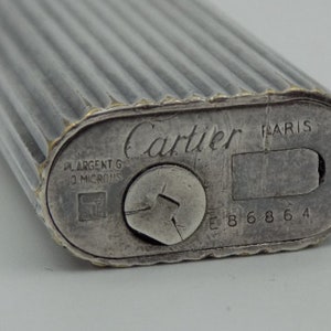 Può includere: Un accendino Cartier in argento con l'iscrizione 'Cartier PARIS' e 'ARGENT G 10 MICRONS' sul fondo. L'accendino ha un design a coste e un numero di serie 'E 8 6 8 6 4'.