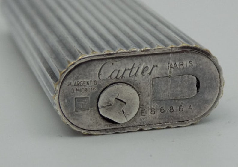 Può includere: Un accendino Cartier in argento con l'iscrizione 'Cartier PARIS' e 'ARGENT G 10 MICRONS' sul fondo. L'accendino ha un design a coste e un numero di serie 'E 8 6 8 6 4'.