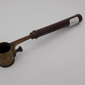 Puede incluir: Una herramienta de medición de latón con un mango de madera. La herramienta tiene una taza redonda con la inscripción "GRAMS PER FLUID OZ 4.0".