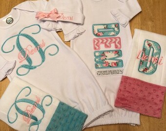May include: A white baby gown with a turquoise and pink floral design, a matching hat, and a white burp cloth with turquoise and pink trim. The gown and burp cloth are embroidered with the name "Demi".