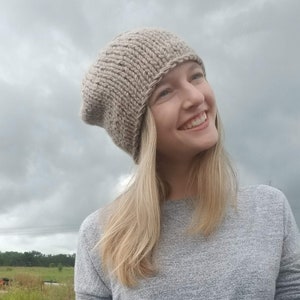 May include: A woman wearing a grey knitted beanie and a grey long-sleeved top. She is standing in a field with a pile of logs behind her.