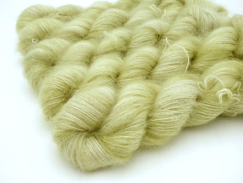 May include: Close-up of several skeins of soft, pale green yarn. The yarn has a fluffy, textured appearance with subtle white highlights. The skeins are arranged closely together, creating a dense, tactile surface. Ideal for knitting and crochet projects.