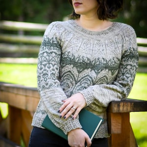 May include: A woman wearing a grey and green patterned jumper with a book in her hands. The jumper has a lace-like design and is knitted with a fine yarn.