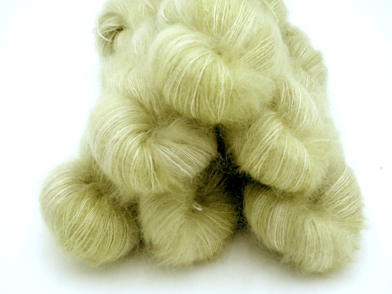 May include: A pile of soft, pale green yarn with a fluffy texture. The yarn appears to be a blend of fibers, with a subtle sheen and white highlights. Ideal for knitting or crocheting projects.
