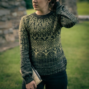 May include: A dark green and gold sweater with a geometric pattern. The sweater is knit and has a loose fit. The person wearing the sweater is holding a book.
