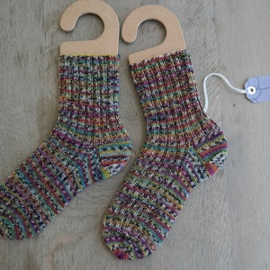 Puede incluir: Un par de calcetines coloridos tejidos a mano con un hilo multicolor, colgados de perchas de cartón. Los calcetines están hechos con una variedad de colores, incluyendo negro, blanco, rojo, amarillo, verde y azul. Los calcetines están etiquetados con una etiqueta que dice "ooh made of wool".