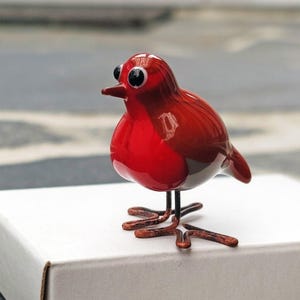 May include: A small, glossy red bird figurine with large black eyes and a small beak. The bird has delicate metal legs and is perched on a white box. The figurine is a decorative item.