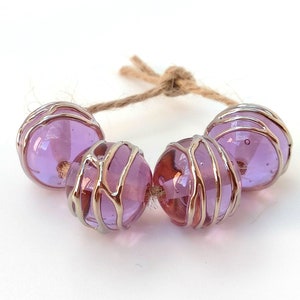 Puede incluir: Una cadena de cuatro cuentas de vidrio translúcido de color lavanda. Cada cuenta está encerrada en una jaula de alambre plateado y ensartada en un cordel natural. Las cuentas son redondas y tienen una cualidad reflectante.