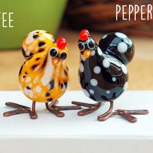 May include: Two glass hen figurines, one in yellow and black with the word "TOFFEE", the other in black and white with the word "PEPPERCORN". Both have red crests and copper wire legs, standing on a white surface.