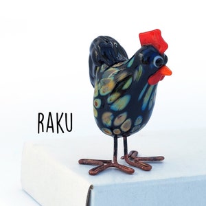 May include: Black and blue glass chicken figurine with a red comb and wattle. The chicken is standing on a white box with the word "RAKU" printed on it.