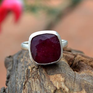 Può includere: Un anello d'argento con una grande gemma di rubino a taglio quadrato. Il rubino è di un colore rosso intenso ed è incastonato in una semplice fascia d'argento lucida. L'anello è esposto su un pezzo di legno.