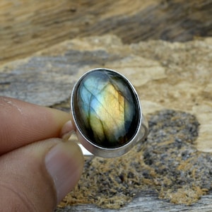 May include: A silver ring featuring a large oval labradorite gemstone. The gemstone displays a green, blue, and gold iridescent sheen.