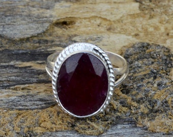 May include: A silver ring with a large, oval-cut, deep red gemstone. The gemstone is set in a decorative bezel with a twisted rope design. The ring is displayed on a textured, light brown wooden surface.