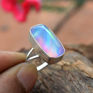 May include: A silver ring featuring a large, rectangular iridescent gemstone. The gemstone displays a range of colors, including blue, purple, and green. The ring is held by a person's fingers, showcasing the ring's design and the play of light on the stone.