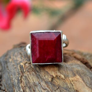 May include: A square-cut, deep red gemstone ring with a silver band. The gemstone is set in a square silver bezel. The ring is displayed on a piece of wood.
