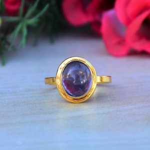 May include: A gold ring with a purple oval gemstone set in a bezel setting. The ring is on a light wood surface.
