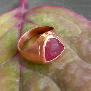 May include: A copper ring with a triangular pink gemstone set in the center. The ring is on a green leaf.