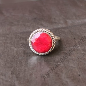 May include: A silver ring with a red gemstone set in a rope-like design. The ring is on a gray textured surface.