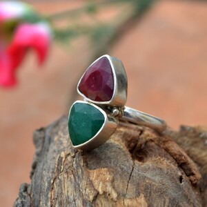 May include: A sterling silver ring featuring two triangular gemstones. The top stone is a deep red, and the bottom stone is a vibrant green. The ring is set on a wooden surface, showcasing the contrast of colors and textures.