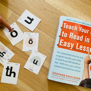 Puede incluir: Un libro titulado "Teach Your Child to Read in 100 Easy Lessons" con tarjetas didácticas con letras y palabras. Las tarjetas didácticas incluyen las letras "th" y "u", y las palabras "orange" y "umbrella". La portada del libro es roja y blanca.