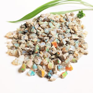 Op de afbeelding: Een grote hoop ruwe, natuurlijke opaal edelstenen in verschillende tinten wit, grijs, blauw, groen en oranje. De opalen zijn verspreid over een wit oppervlak.