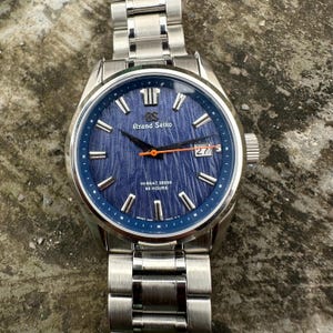 May include: A stainless steel Grand Seiko watch with a textured blue dial, silver indices, and a bright orange second hand. The dial features the text Grand Seiko, HI-BEAT 36000, and 80 HOURS, with a date window at the three o'clock position.