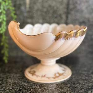 May include: A vintage, footed bowl with a scalloped rim and a spiral design. The bowl is a pale peach color with gold trim along the rim. The base of the bowl has a decorative gold floral pattern.