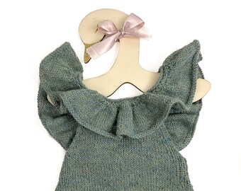 May include: A green knitted baby onesie with a ruffled neckline and short sleeves. The onesie is hanging on a wooden swan-shaped hanger with a pink bow.