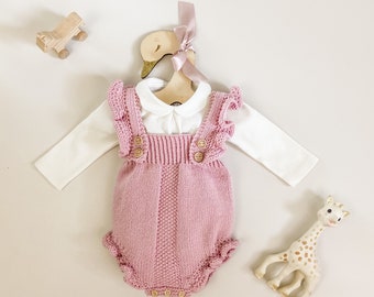 May include: A pink knitted baby romper with ruffle sleeves and wooden button closures. The romper is paired with a white long-sleeve shirt with a peter pan collar.