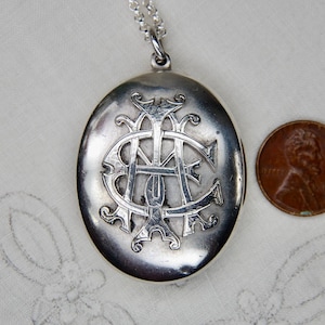 May include: Oval silver locket pendant with an ornate monogram, suspended from a delicate chain. A penny is placed next to the locket for size comparison. The locket has a detailed, engraved design.