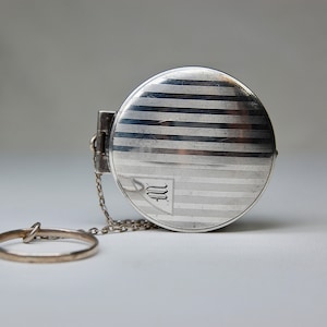 May include: A silver compact with a chain and a monogrammed "M" on the front. The compact has a striped design.