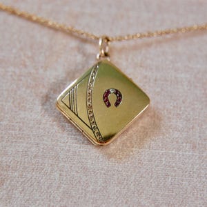 Könnte beinhalten: Goldfarbene Halskette mit quadratischem Medaillon. Das Medaillon zeigt ein Hufeisen-Design mit roten Akzenten und dekorativen Linien. Die Halskette hat eine feine Goldkette.