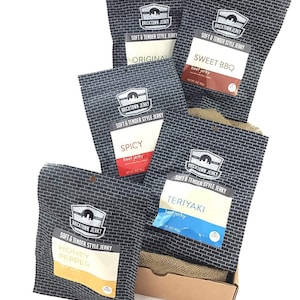 May include: A gift box filled with five different flavors of beef jerky. The flavors are Original, Sweet BBQ, Spicy, Teriyaki, and Honey Pepper. Each bag is black with white text and a brick pattern.