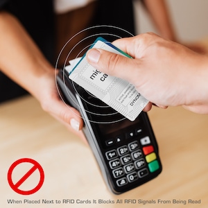May include: A white and blue card labeled "mic ca" is held over a black card reader. The card has a circuit board design and the text "ACTIVE JAMMING TI Anti-Theft RFID Block 13.56 Mhz DYNOM". The image shows a payment process.