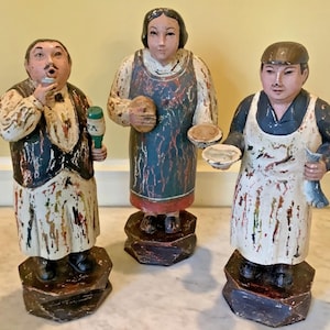May include: Three hand-painted wooden figurines standing on hexagonal bases. The left figure wears a vest and apron, holding a bottle. The center figure wears a blue apron and holds a round object. The right figure wears an apron and holds a fish.