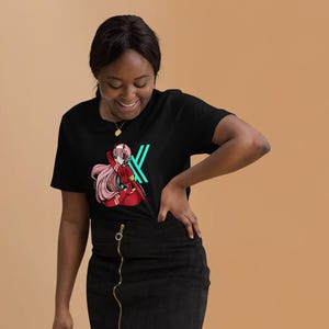 May include: Black t-shirt with a colourful anime-style graphic and a teal and white letter design. The person is wearing a black skirt and white trainers. A black purse is held in the left hand.