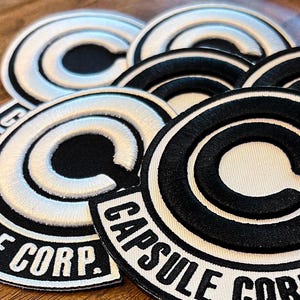 May include: Circular black and white embroidered patches with the text "CAPSULE CORP." These patches feature a stylized "C" design. The patches are arranged on a wooden surface.
