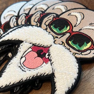 Puede incluir: Parches bordados con una calavera de dibujos animados que lleva gafas de sol rojas con lentes verdes. Los parches tienen un fondo beige y costuras negras. Un parche muestra una criatura blanca y esponjosa con boca rosa y contorno negro.