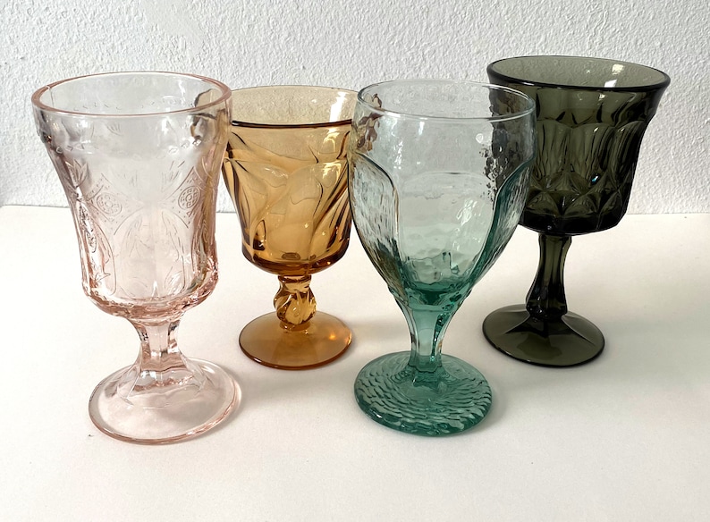 May include: Four vintage stemmed glasses in different colours and patterns. The glasses are pink, amber, green, and grey.