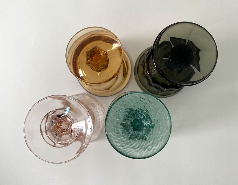 May include: Four vintage glass drinking glasses with different colours and textures. The glasses are clear, pink, green, and brown.