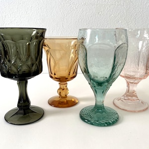 May include: Four stemmed glasses in different colours and patterns. The glasses are clear, amber, green, and pink.