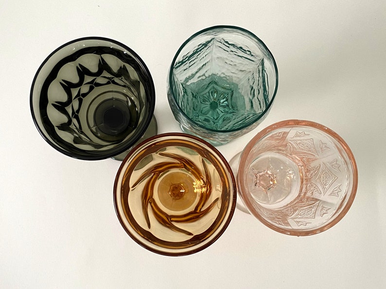 May include: Four vintage drinking glasses with different patterns. The glasses are clear, green, brown, and pink.