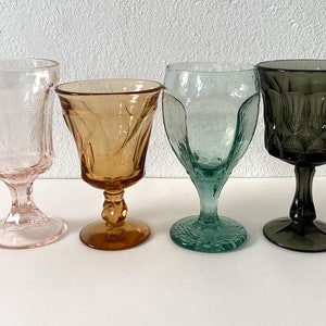 May include: Four stemmed glasses in different colours: pink, amber, green, and grey. The glasses are all similar in shape and have a textured design.