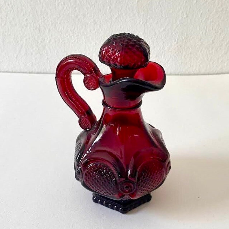Puede incluir: Una botella de vidrio rojo rub&iacute; con una superficie texturizada y un tap&oacute;n decorativo. La botella tiene un asa y un pico para verter.