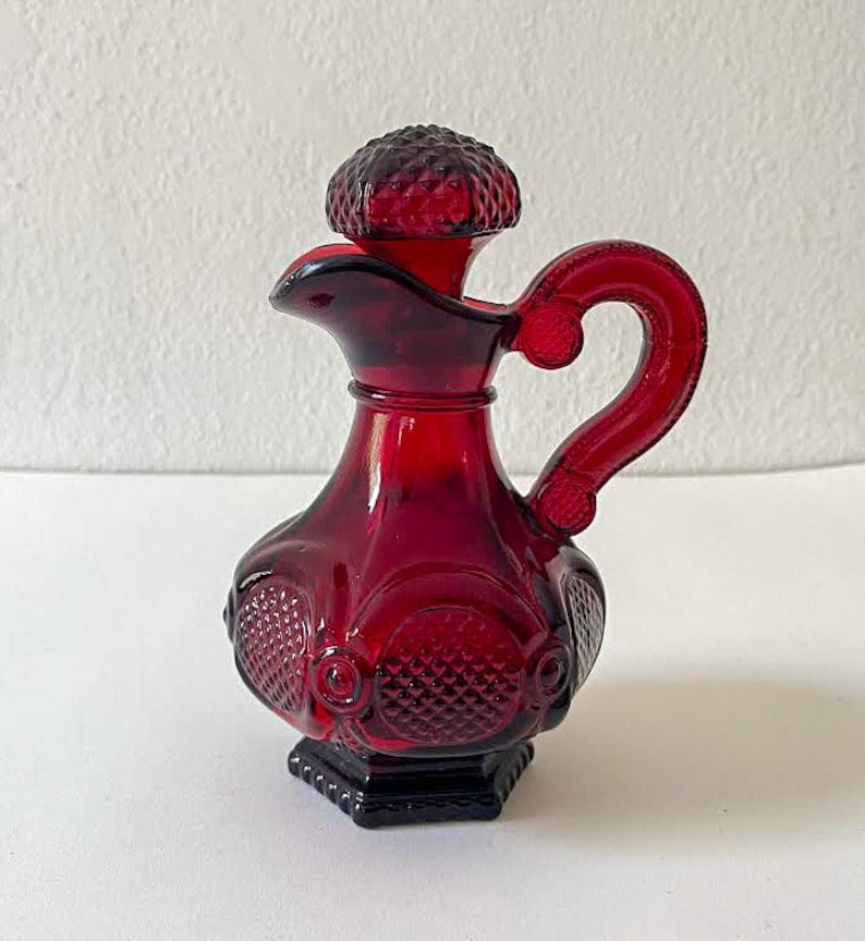 Puede incluir: Una botella de vidrio roja con un tap&oacute;n decorativo y un asa. La botella tiene un patr&oacute;n de diamante y un dise&ntilde;o en relieve.